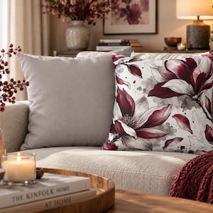 May include: A square throw pillow featuring a watercolour-style floral print with deep burgundy and white magnolia blossoms against a white background with soft grey accents, paired with a solid light grey square throw pillow.