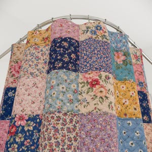 May include: A patchwork shower curtain with a floral pattern in various colors, including pink, blue, yellow, and purple. The curtain is quilted and hangs from a curved metal rod, adding a touch of vintage charm to any bathroom.