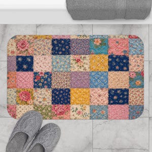 May include: A rectangular bath mat with a patchwork design. The mat features a variety of floral patterns in pink, blue, yellow, and purple squares. Gray slippers are at the bottom left corner. A gray towel is at the top.