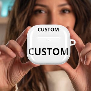 May include: A white, square-shaped case with the word "CUSTOM" printed in black on the top and bottom. The case has a rounded top and a small loop on the side. The background is blurred, with a person holding the case.