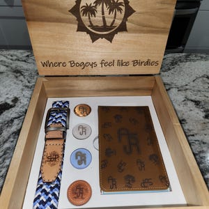 May include: A wooden box with a lid that opens to reveal a golf themed gift set. The box is engraved with the words "Where Bogeys feel like Birdies" and a sun with palm trees. The gift set includes a blue and white patterned belt, a brown leather golf scorecard holder, and four golf ball markers.