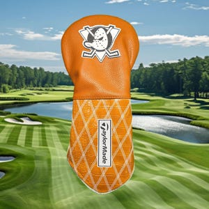 Könnte beinhalten: Ein orangefarbener Golfschlägerhaube mit einer strukturierten, lederartigen Oberfläche. Der obere Teil zeigt ein weiß-schwarzes Entenlogo mit gekreuzten Hockeyschlägern. Der untere Teil hat ein weißes Rautenmuster und ein vertikales TaylorMade-Logo.