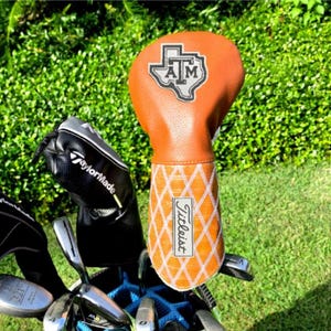 Puede incluir: Una funda para palo de golf de color naranja con un patrón de diamantes blancos y el logotipo de Titleist. La parte superior presenta un parche con forma de Texas con las letras ATM. Se muestra junto a fundas negras TaylorMade y palos de golf.