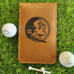 May include: A brown leather golf scorecard holder with a Seminole State University logo. The logo is a black and white image of a Native American man's head with a feather headdress. The holder is on a green grass background with two white golf tees and two white golf balls.