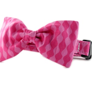 May include: A pink bow tie with a wavy pattern, attached to a black adjustable collar.