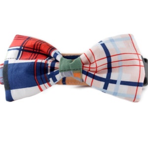 May include: A colorful plaid bow tie with red, blue, white, and pink squares. The bow tie is made of fabric and has a brown leather strap.