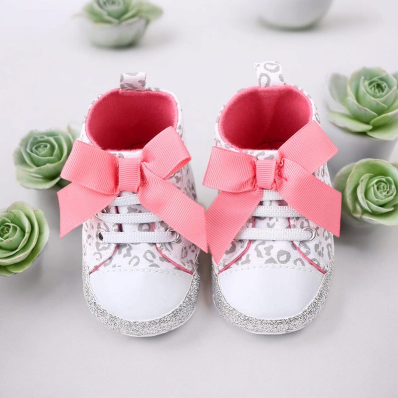 May include: A pair of white baby shoes with a pink bow and a leopard print design. The shoes have silver glitter on the bottom.