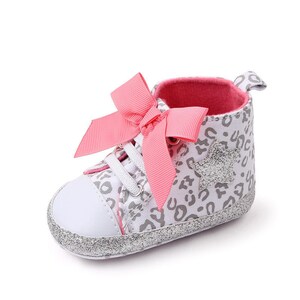 May include: A pair of baby shoes with a white canvas upper featuring a grey leopard print and a silver star. The shoes have pink satin bows and a silver glitter trim.