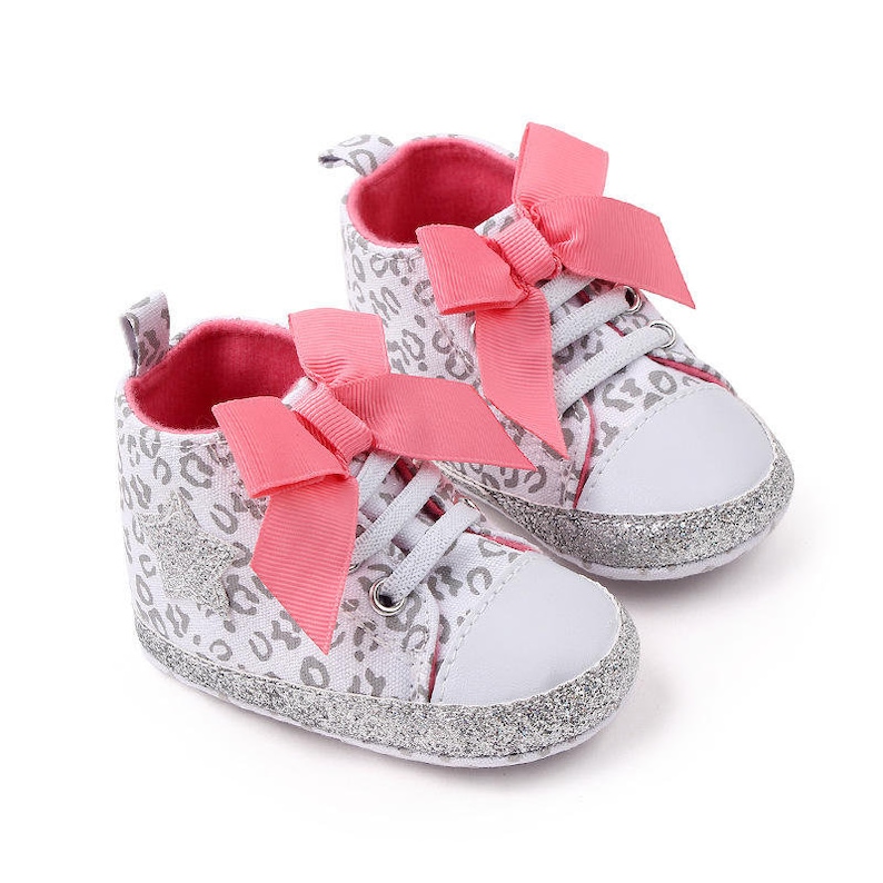May include: A pair of baby shoes with a white canvas upper and a silver glitter trim. The shoes have a pink bow on the front and a leopard print design.