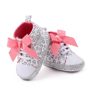 May include: A pair of white baby shoes with a grey leopard print and pink bows. The shoes have silver glitter accents.