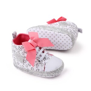 May include: A pair of white baby shoes with a silver glitter finish. The shoes have a pink bow and a leopard print design.