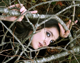 May include: A woman with dark hair and dark eye makeup is lying in a tangle of branches. Her eyes are looking directly at the camera.