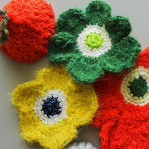 May include: Close-up of handmade crochet flowers in vibrant colors. The arrangement includes a red apple-shaped item with a green stem, and several flowers in green, yellow, and red, each with contrasting centers.