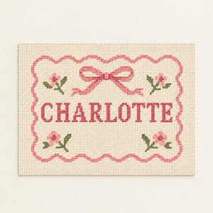 May include: A rectangular cross-stitch embroidery on cream-colored fabric featuring the name CHARLOTTE in pink capital letters. The design includes a pink scalloped border, a central pink bow, and four small pink flowers with green leaves.