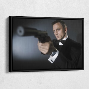 May include: A man in a black tuxedo and bow tie holds a gun in his right hand, aiming it towards the camera. The background is a dark gray.