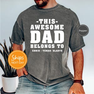 May include: A gray t-shirt with the text "-THIS- AWESOME DAD BELONGS TO CHRIS - YENAH - GLADYS" in white block letters. The shirt has a vintage, washed look.