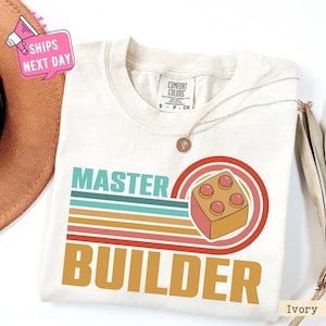 May include: Ivory t-shirt with a retro-style graphic that says "Master Builder" with a colorful striped background and a cartoon image of a red and yellow Lego brick.