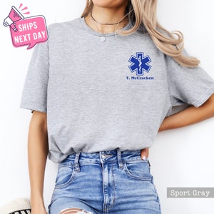 May include: A gray t-shirt with a blue medical star of life symbol and the text "T. McCracken" printed on the front.