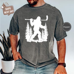 May include: A gray t-shirt with a white graphic of a Bigfoot holding a hockey stick and standing in a forest. The text "Pepper" is in the upper right corner of the image.