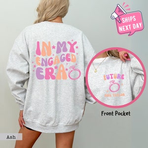 May include: Ash-colored sweatshirt with the text "IN MY ENGAGED ERA" in pink, orange, and purple. A ring graphic is below the text. A smaller image shows "FUTURE" and a ring graphic. The text "MRS. TAYLOR" is below the ring.
