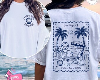 May include: White t-shirt with a blue graphic of a seashell and the text "Katie's Bach San Diego" on the front. The back of the shirt has a blue graphic of a beach scene with palm trees, a beach umbrella, a lounge chair, and the text "San Diego, CA Katie's Bach, 2025".