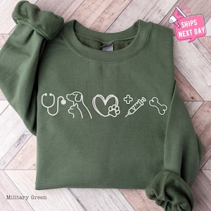 May include: A military green sweatshirt with a white graphic design of a stethoscope, a dog, a cat, a heart with a paw print, a syringe, and a bone. The graphic design is centered on the sweatshirt.