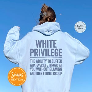 A person wearing a light blue hoodie with the text "WHITE PRIVILEGE" printed on the back, standing against a clear blue sky.