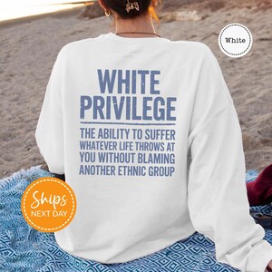 A person wearing a white sweatshirt with the text "WHITE PRIVILEGE" printed on it, sitting on a beach.