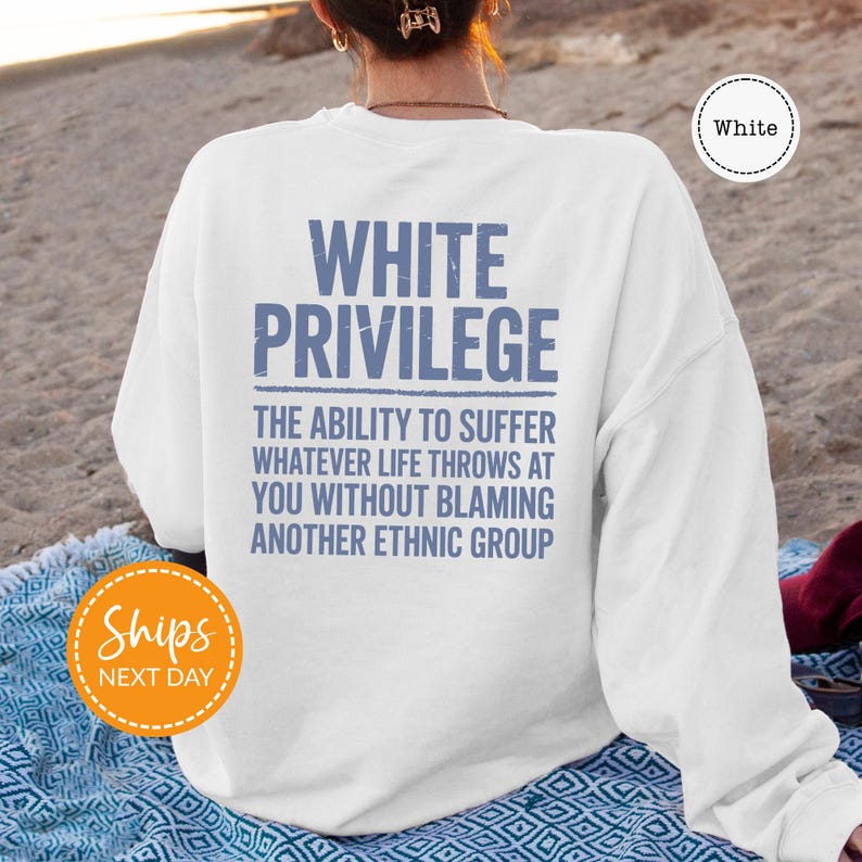 A person wearing a white sweatshirt with the text "WHITE PRIVILEGE" printed on it, sitting on a beach.