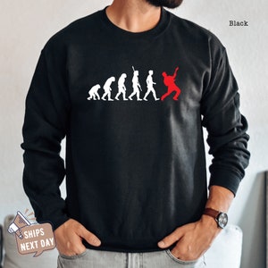 May include: Black crewneck sweatshirt with a white evolution graphic and a red figure playing a guitar. The word "Black" is printed in the upper right corner.