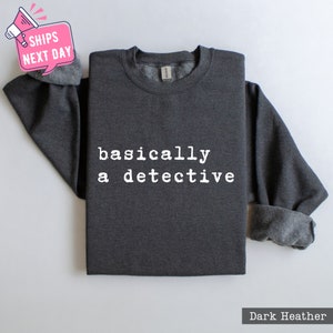 May include: Dark heather grey sweatshirt with the text "basically a detective" printed in white.