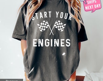 Puede incluir: Camiseta de color pimienta con la frase "START YOUR ENGINES" y dos banderas a cuadros blancas cruzadas. La camiseta tiene un corte holgado y mangas cortas.