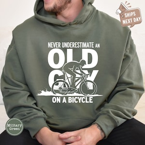 May include: Military green hoodie with the text "NEVER UNDERESTIMATE AN OLD GUY ON A BICYCLE" in white. The graphic shows a cyclist on a bicycle. The hoodie is a comfortable, casual style.
