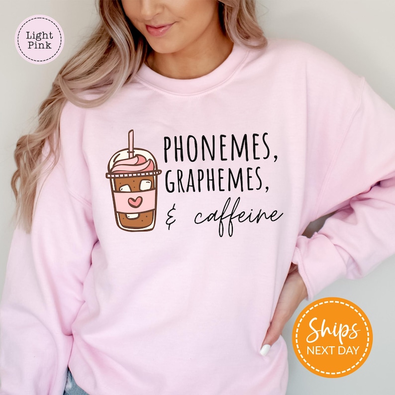 A woman wearing a pink sweatshirt with a graphic design of a coffee cup and text that says "PHONEMES, GRAPHHEMS, & caffeine".