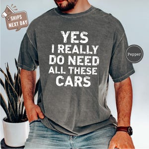 May include: Gray t-shirt with the text "YES I REALLY DO NEED ALL THESE CARS" in white block letters. The shirt is a casual style, suitable for everyday wear. The image also includes a potted plant.