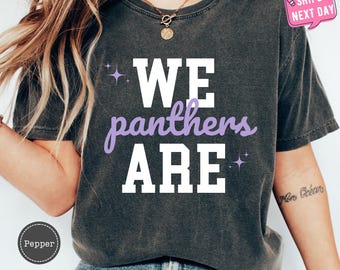 May include: A dark gray t-shirt with the text "WE panthers ARE" in white and purple lettering. The shirt has a distressed look and features small glitter accents.