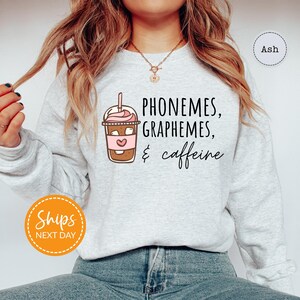 A woman wearing a gray sweatshirt with a graphic design of a coffee drink and text that says "PHONEMES, GRAPHEMES, & caffeine".