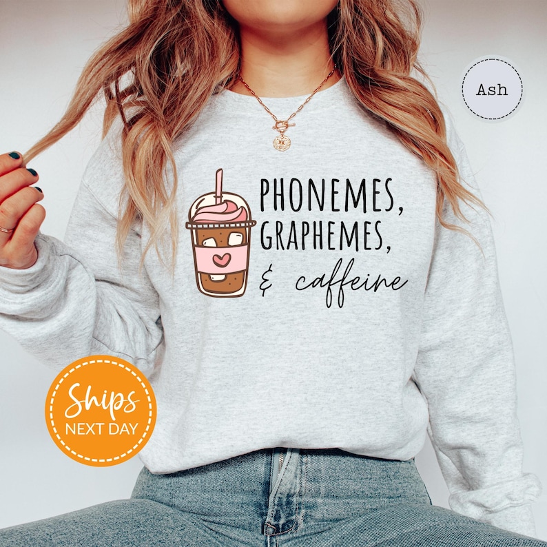 A woman wearing a gray sweatshirt with a graphic design of a coffee drink and text that says "PHONEMES, GRAPHEMES, & caffeine".