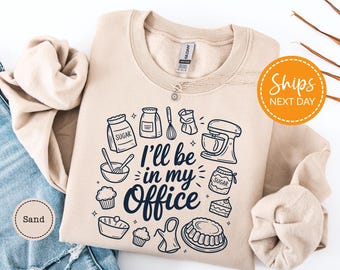 May include: A sand-colored sweatshirt with a black graphic of baking tools and the text "I'll be in my Office." The design includes images of sugar bags, a mixer, and cupcakes. The sweatshirt is folded on a white surface.