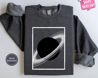 May include: Dark heather grey sweatshirt with a white graphic of a planet with rings. The graphic is framed in a white rectangle.