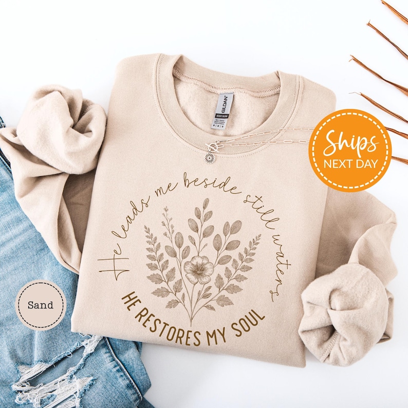 a beige sweatshirt with a floral design and text, along with a pair of blue jeans and some twigs, all laid out on a white background.