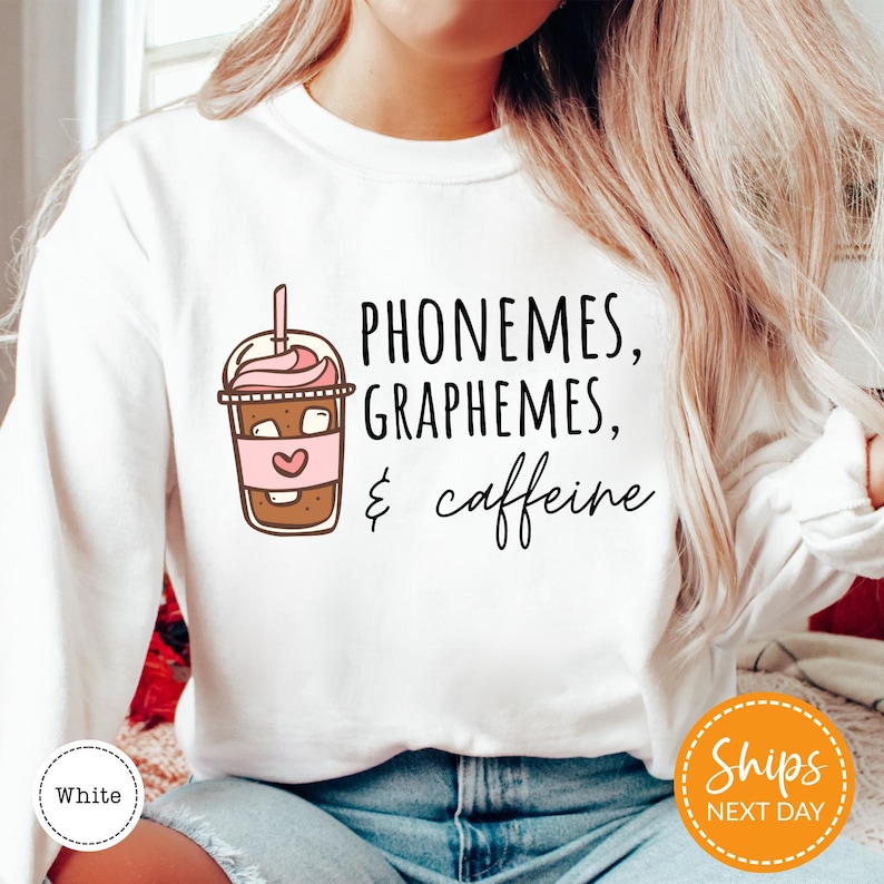A woman wearing a white sweatshirt with a graphic design that says "PHONEMES, GRAPHEMES, & caffeine" printed on it.