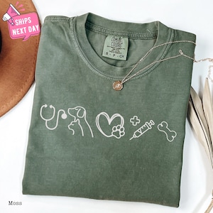 May include: A moss green t-shirt with a white graphic of a stethoscope, a dog, a cat, a heart with a paw print, a plus sign, a syringe, and a bone. The text "COMFORT COLORS" is visible on the tag.