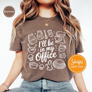 May include: Espresso-colored t-shirt with a white graphic design featuring baking-related illustrations and the text "I'll be in my Office." The design includes images of sugar bags, a mixer, cupcakes, and other baking tools.