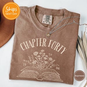 May include: Espresso-colored t-shirt with the text "CHAPTER FORTY" above an open book graphic with flowers. The shirt has a crew neck and short sleeves. A silver necklace with a pendant is draped across the shirt.