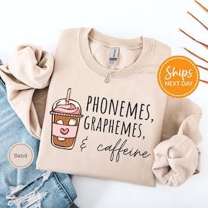 a beige sweatshirt with a graphic design of a coffee drink and the text "Phone me, graph me, caffeine me" printed on it. The sweatshirt is laid out on a white background, along with a pair of blue jeans.