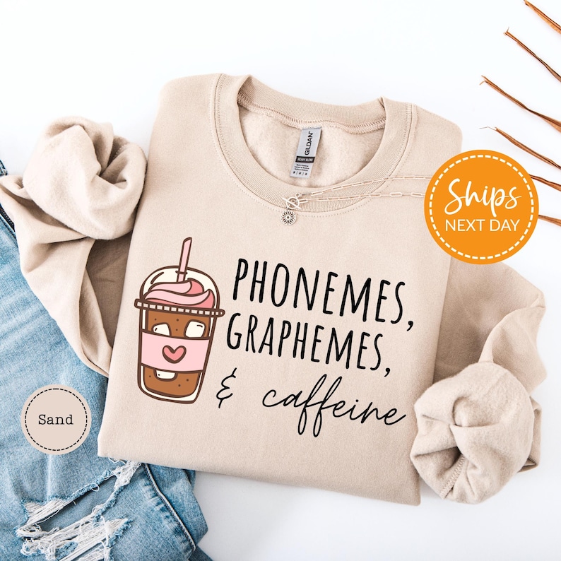 a beige sweatshirt with a graphic design of a coffee drink and the text "Phone me, graph me, caffeine me" printed on it. The sweatshirt is laid out on a white background, along with a pair of blue jeans.