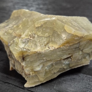 May include: A rough-hewn, pale yellow-green stone with a layered, textured appearance. The natural mineral displays variations in color and surface detail, with hints of brown and gray. The stone is likely a raw, unpolished specimen.