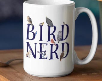 May include: A white ceramic mug featuring the dark blue text BIRD NERD. The design includes various small, detailed illustrations of different bird species perched on and around the letters, including owls, a cardinal, and a bald eagle.