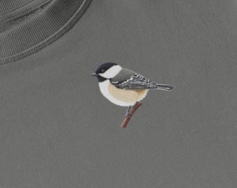 May include: A gray sweatshirt featuring an embroidered chickadee bird perched on a brown branch. The bird has a black and white head, gray wings, and a cream-colored belly. The sweatshirt is a solid, muted gray color.